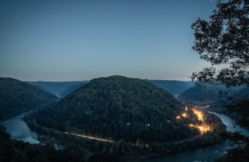 A serene landscape at dusk featuring a river winding around a forested hill, with lights from a nearby town.