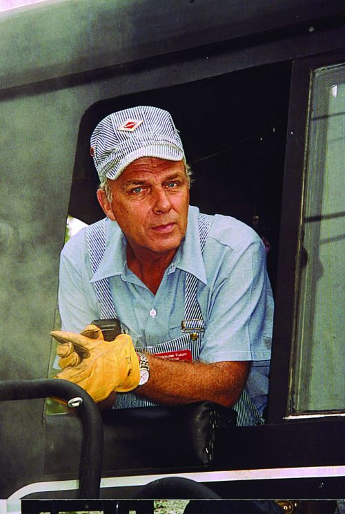 A man in a striped cap and gloves leans out of a train cab, surrounded by steam, looking directly at the camera.