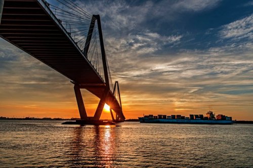 A large bridge spans over water at sunset, with a cargo ship passing beneath and clouds in the sky.