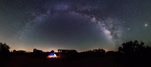 A tent illuminated by a warm glow under a starry sky with the Milky Way arching overhead.