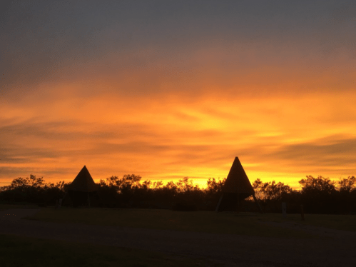 Silhouettes of two triangular structures against a vibrant orange and yellow sunset sky.