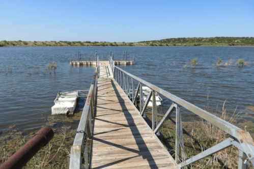 A wooden dock extends over calm water, leading to a small platform surrounded by greenery and blue sky.