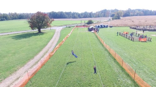 Aerial view of a zip line in a grassy field with people and orange fencing in the background.