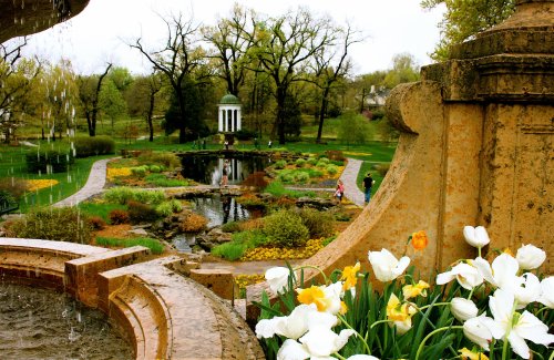 A scenic garden with a fountain, colorful flowers, and a gazebo surrounded by lush greenery and walking paths.