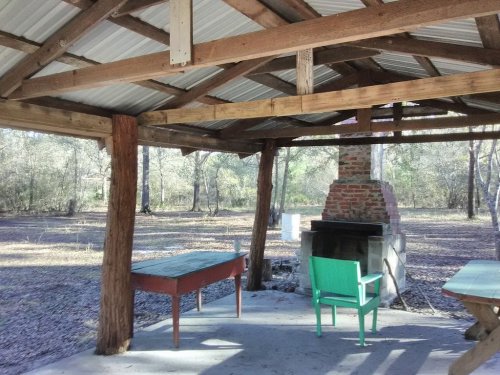 A rustic outdoor shelter with wooden beams, a stone fireplace, and green and red furniture in a wooded area.