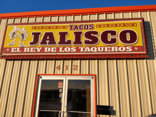 Sign for "Tacos Jalisco - El Rey de los Taqueros" on a colorful storefront, featuring vibrant lettering and a decorative design.