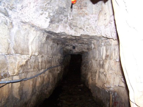 A dimly lit underground tunnel with stone walls and a narrow pathway, featuring exposed electrical wires.
