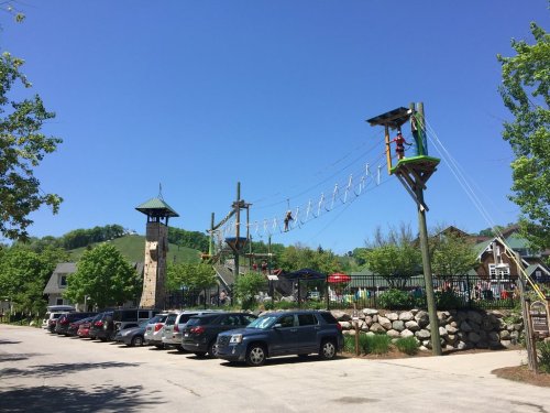 A sunny day at an adventure park with a ropes course, surrounded by trees and parked cars in the foreground.