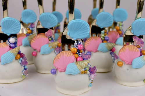 Decorative cake pops topped with colorful seashells and beads, each held by a gold fork.