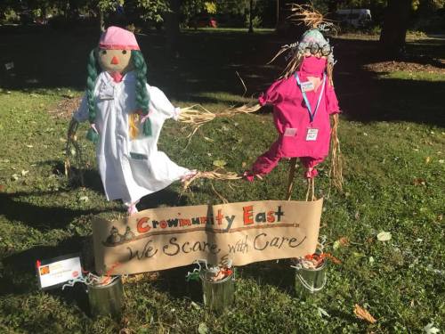 Two scarecrows in medical attire stand beside a sign that reads "Crownmunity East: We Scare with Care."