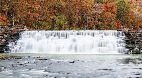 A serene waterfall cascades over stone steps, surrounded by vibrant autumn foliage and a calm river.