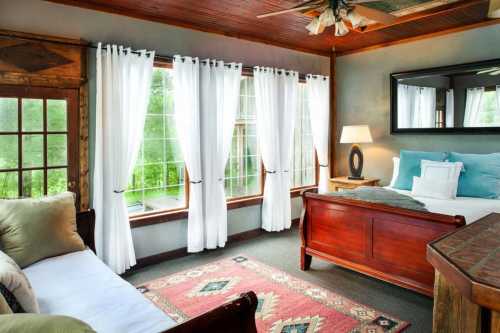 Bright, cozy bedroom with large windows, white curtains, a wooden bed, and a decorative rug.