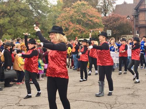 A group of dancers in red sequined tops and black hats perform in a lively parade, surrounded by colorful spectators.