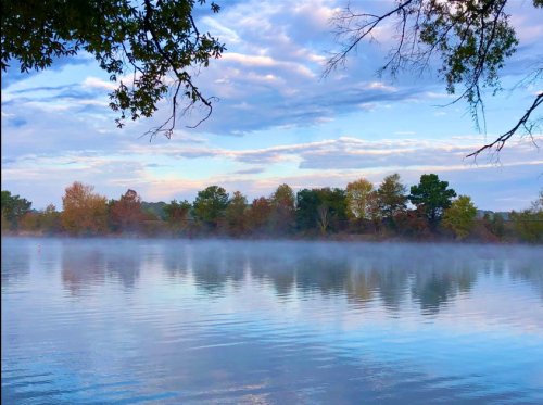 A serene river scene at dawn, with mist rising from the water and trees reflecting on the calm surface.