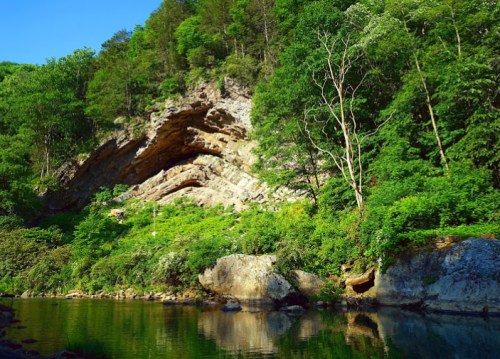 Lush green trees and rocky cliffs reflect in a calm river under a clear blue sky.