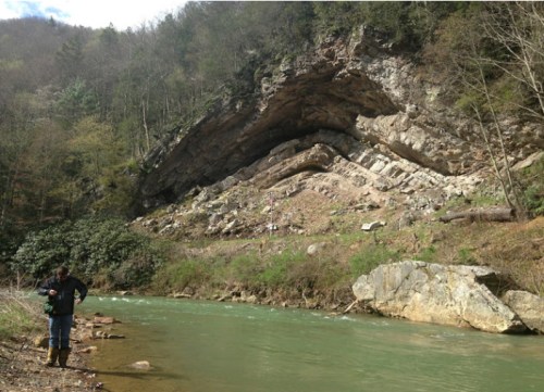 A person stands by a riverbank, with a rocky cliff and trees in the background. The water is a light green color.