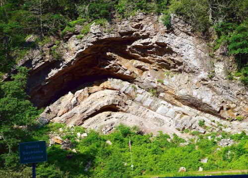 Rock formation with layered, curved strata surrounded by greenery, near a sign for Devil's Backbone Anticline.