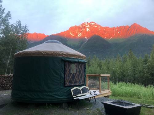 A green yurt sits in a scenic landscape, with mountains bathed in sunset light and trees surrounding the area.
