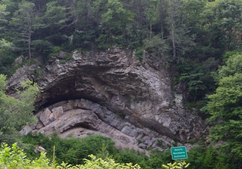 A rocky hillside with layered formations and dense green trees, featuring a sign for "Devils Backbone Anticline."