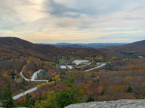 A panoramic view of a valley with winding roads and autumn foliage, surrounded by mountains under a cloudy sky.
