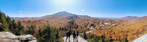 A panoramic view of autumn foliage with two people standing on a rocky ledge overlooking a valley and mountains.