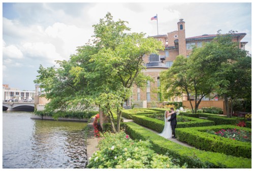 A couple stands together in a lush garden by a river, with a historic building and American flag in the background.