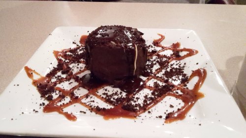 A chocolate dessert on a white plate, drizzled with caramel and surrounded by crushed cookies.