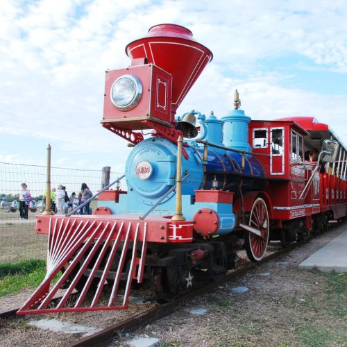 A colorful vintage steam train with a red and blue engine, parked on a track with people in the background.