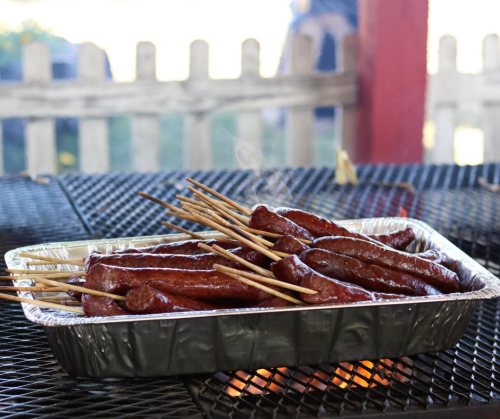 A tray of grilled sausages on skewers, resting on a grill with smoke rising in the background.