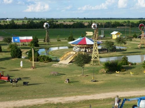 A scenic view of a park with a gazebo, pond, windmills, and a Texas flag, surrounded by green fields and blue skies.