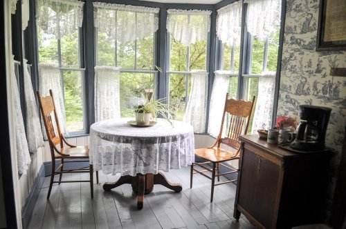 A cozy sunlit room with a round table, two wooden chairs, and a coffee maker, surrounded by lace curtains and greenery.