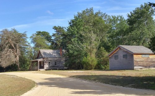 Two rustic wooden buildings surrounded by trees, with a sandy path leading towards them under a clear blue sky.
