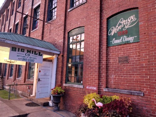 Exterior of a brick building with signs for "Big Mill Co." and "Vintage 1889," featuring colorful plants in front.