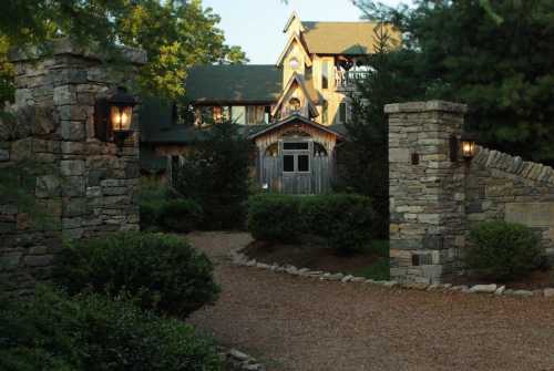 A stone entrance with lanterns leads to a rustic house surrounded by trees and greenery.