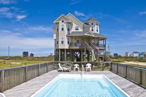 A beach house on stilts with a pool in the foreground, surrounded by grassy land and a clear blue sky.