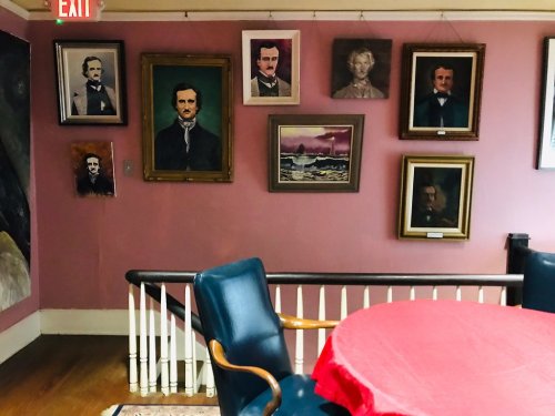 A pink wall adorned with various framed portraits, including historical figures, beside a round table and chairs.