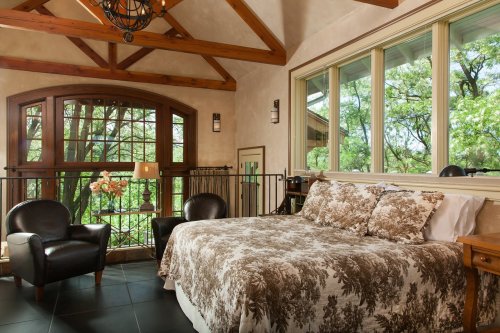 Cozy bedroom with a large bed, two chairs, and large windows overlooking greenery, featuring wooden beams and warm decor.