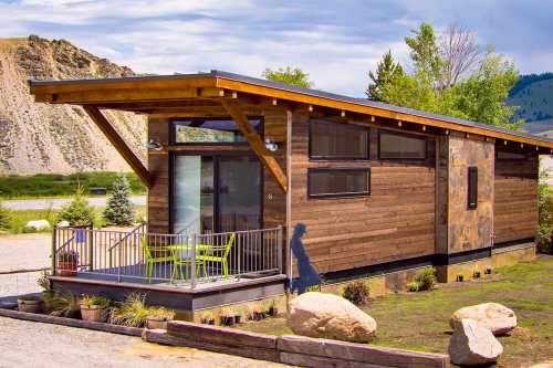 A modern wooden tiny house with large windows, a deck, and green chairs, set in a scenic outdoor landscape.