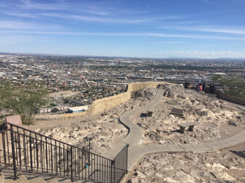 A rocky overlook with a winding path, offering a panoramic view of a cityscape under a clear blue sky.
