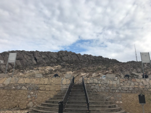 Stone steps lead up to a rocky hillside under a cloudy sky, with a few signs visible along the path.