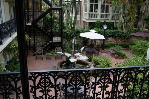 A serene courtyard with a fountain, surrounded by greenery and seating, viewed from a balcony with ornate ironwork.