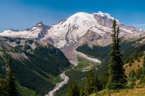 A majestic snow-capped mountain rises above a lush green valley, with a winding river and clear blue sky.