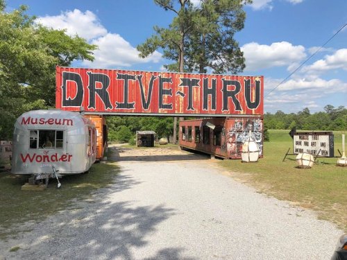A colorful "DRIVE THRU" sign welcomes visitors to a quirky roadside attraction featuring a vintage trailer.