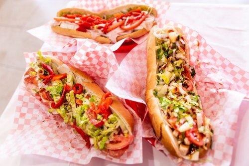 Three colorful sandwiches on red and white checkered paper, filled with fresh vegetables and meats.