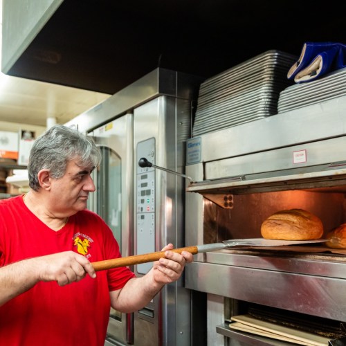 A baker in a red shirt uses a wooden paddle to check bread in a commercial oven.