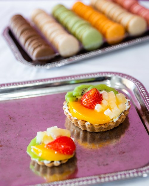 A silver tray with two fruit tarts in the foreground and a colorful assortment of macarons in the background.