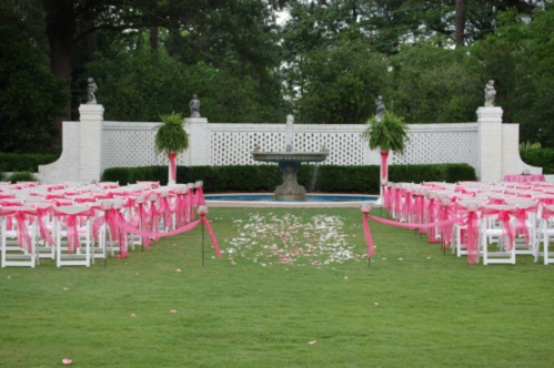 A wedding setup with white chairs adorned with pink ribbons, leading to a fountain surrounded by greenery and flower petals.