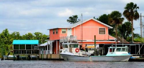 A pink house by the water with boats docked nearby, surrounded by trees and a cloudy sky.