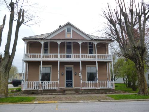 A two-story house with a porch, surrounded by trees, featuring a light brown exterior and a blue front door.