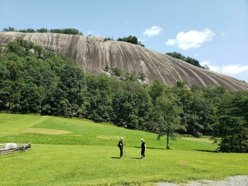 Two people stand on a grassy field in front of a large, smooth rock formation surrounded by trees and blue sky.